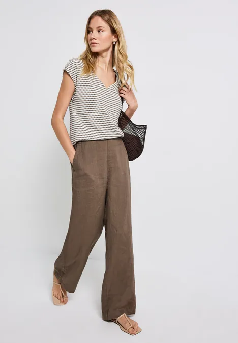 Frau in gestreiftem Shirt, weiter Hose, Sandalen und Netz-Tasche vor weißem Hintergrund.